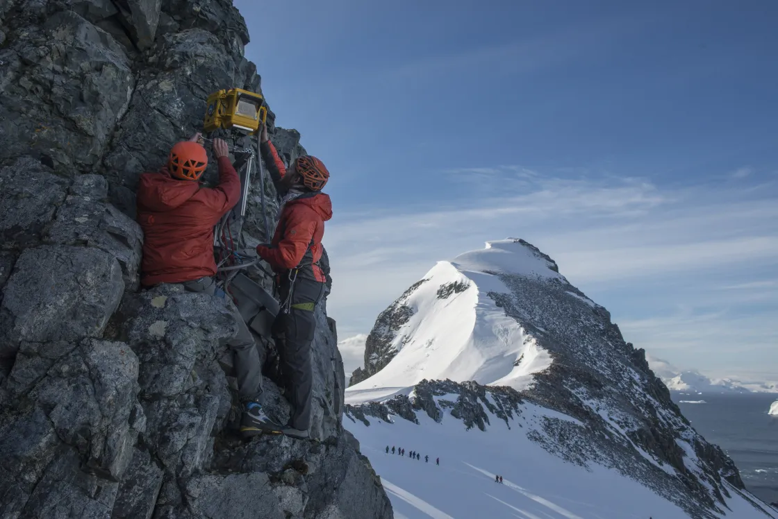 Matthew Kennedy and Daniel McGrath of the Extreme Ice Survey team install a camera at Orne Harbor on the Antarctica Peninsula. In the background, hikers are visible from the National Geographic Explorer, the ship that brought the Extreme Ice Survey team to its sites on the Antarctic Peninsula and South Georgia.